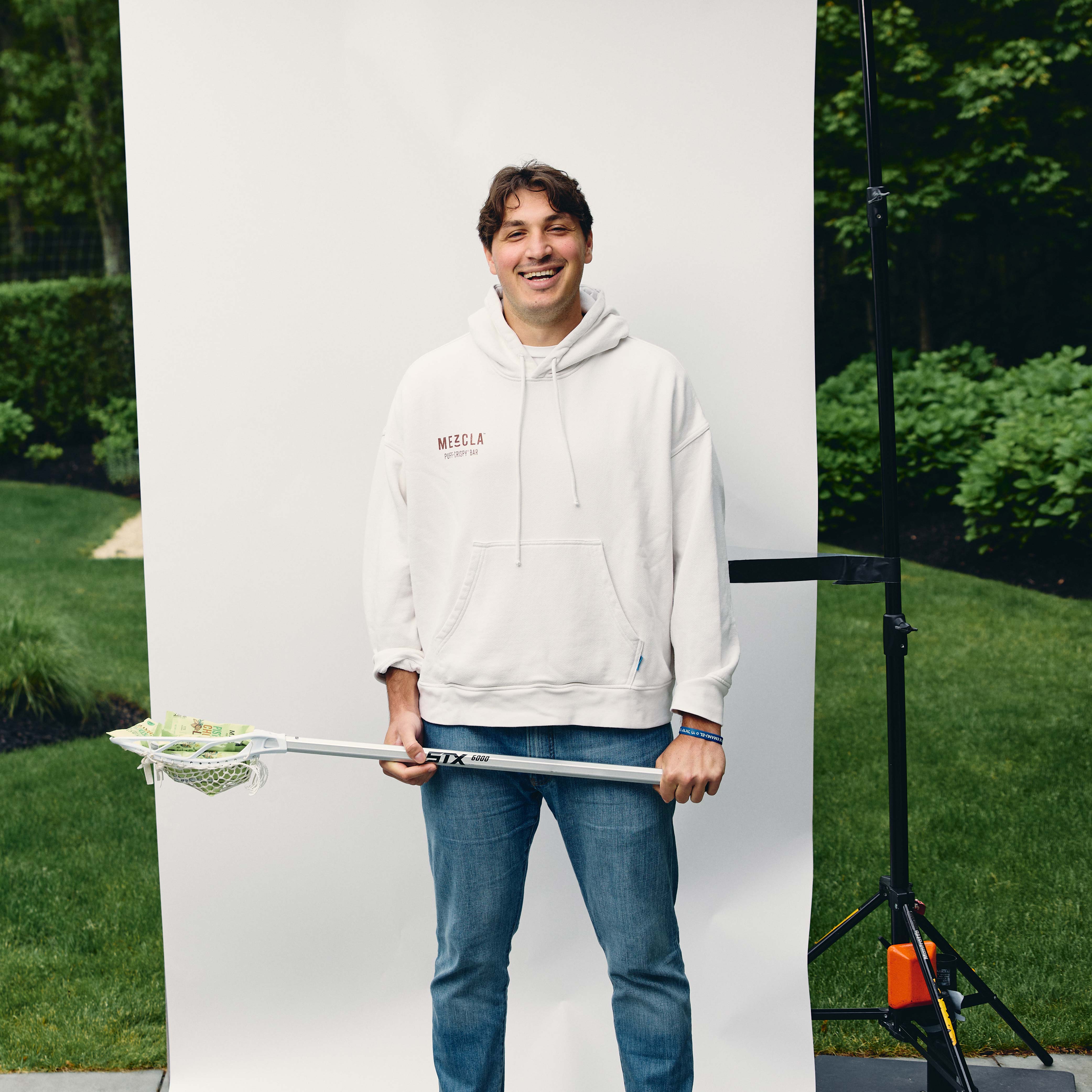 Man in white Mezcla hoodie and jeans smiling while holding lacrosse stick, standing against white backdrop outdoors
