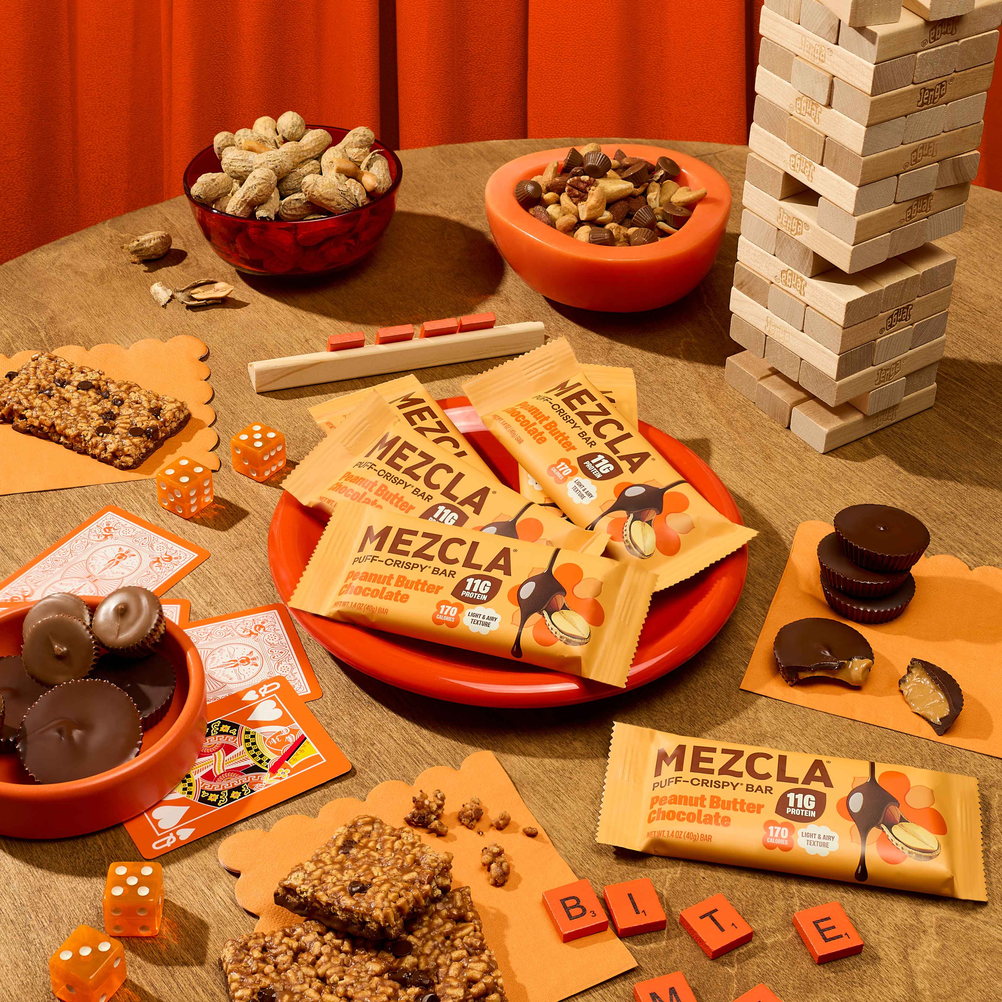 Mezcla protein bars displayed on wooden surface with orange bowls of snacks, dice, cards, Jenga blocks, and chocolate treats in warm orange-themed setting