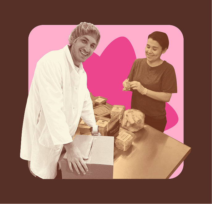 Two friends in a pink-framed scene: one in white chef coat smiling, another in brown shirt, both handling small boxes on a wooden table with a dog nearby.