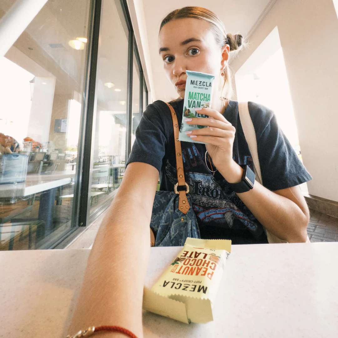 Young woman with blonde hair in bun drinking matcha vanilla drink from white Mezcla package in bright modern cafe setting