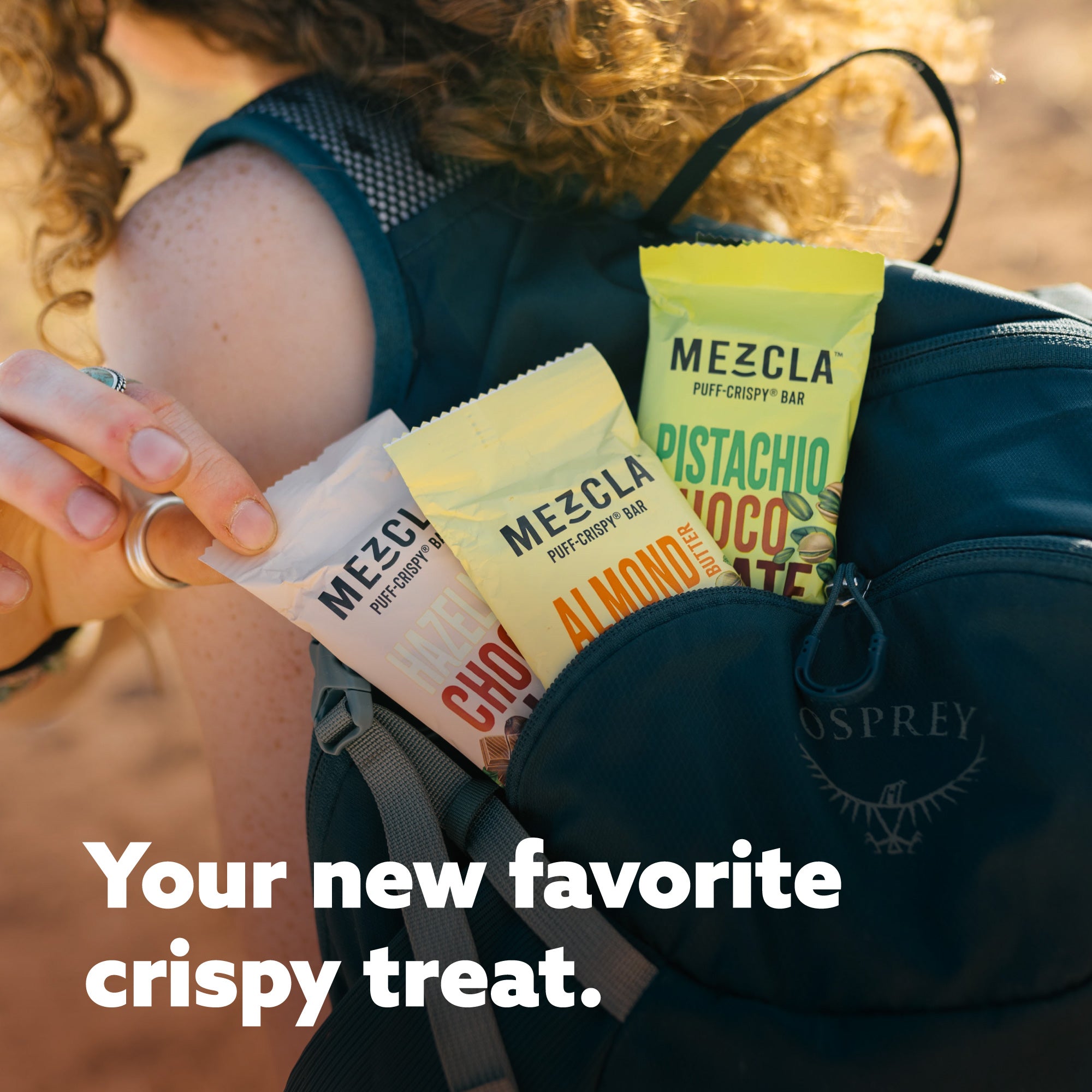 Woman in teal athletic wear holding colorful Mezcla puff-crispy bar packages near Osprey backpack with text "Your new favorite crispy treat"