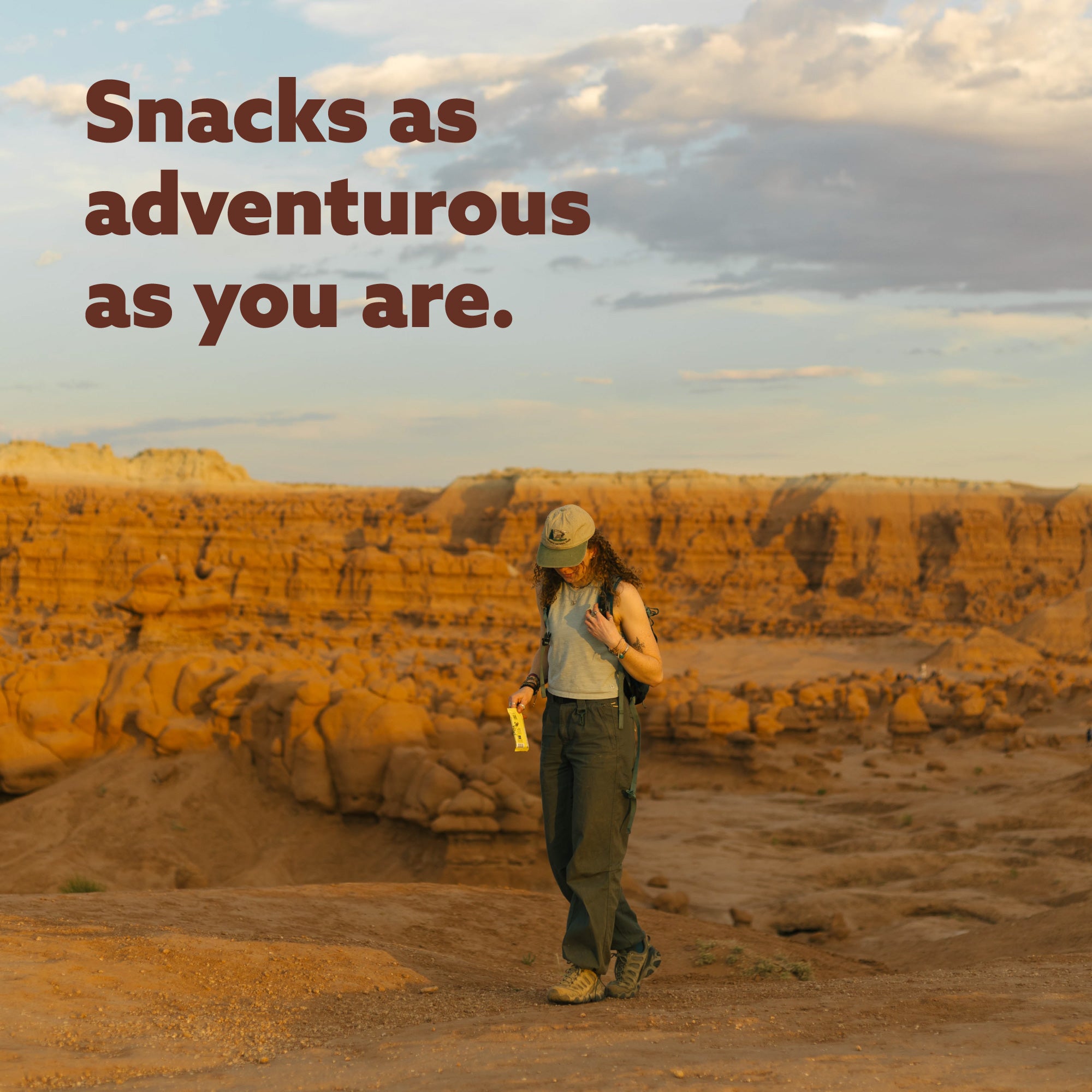 Person in desert landscape holding yellow snack package with text "Snacks as adventurous as you are" overlaid on canyon scenery