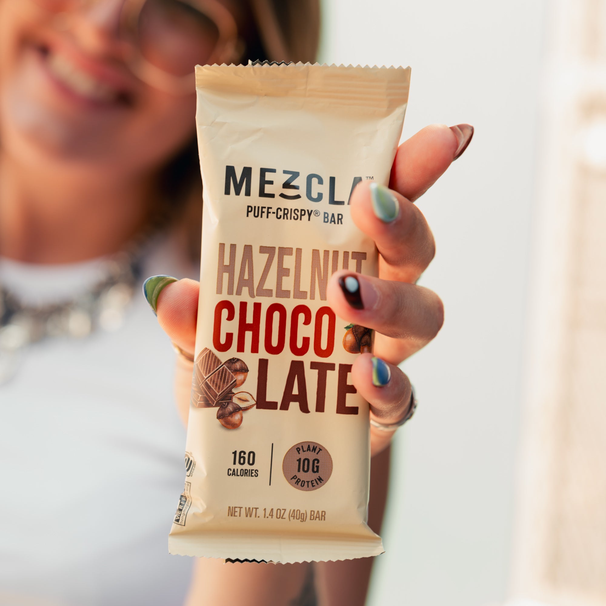 Hand with colorful nail polish holding Mezcla Hazelnut Chocolate protein bar package against blurred background