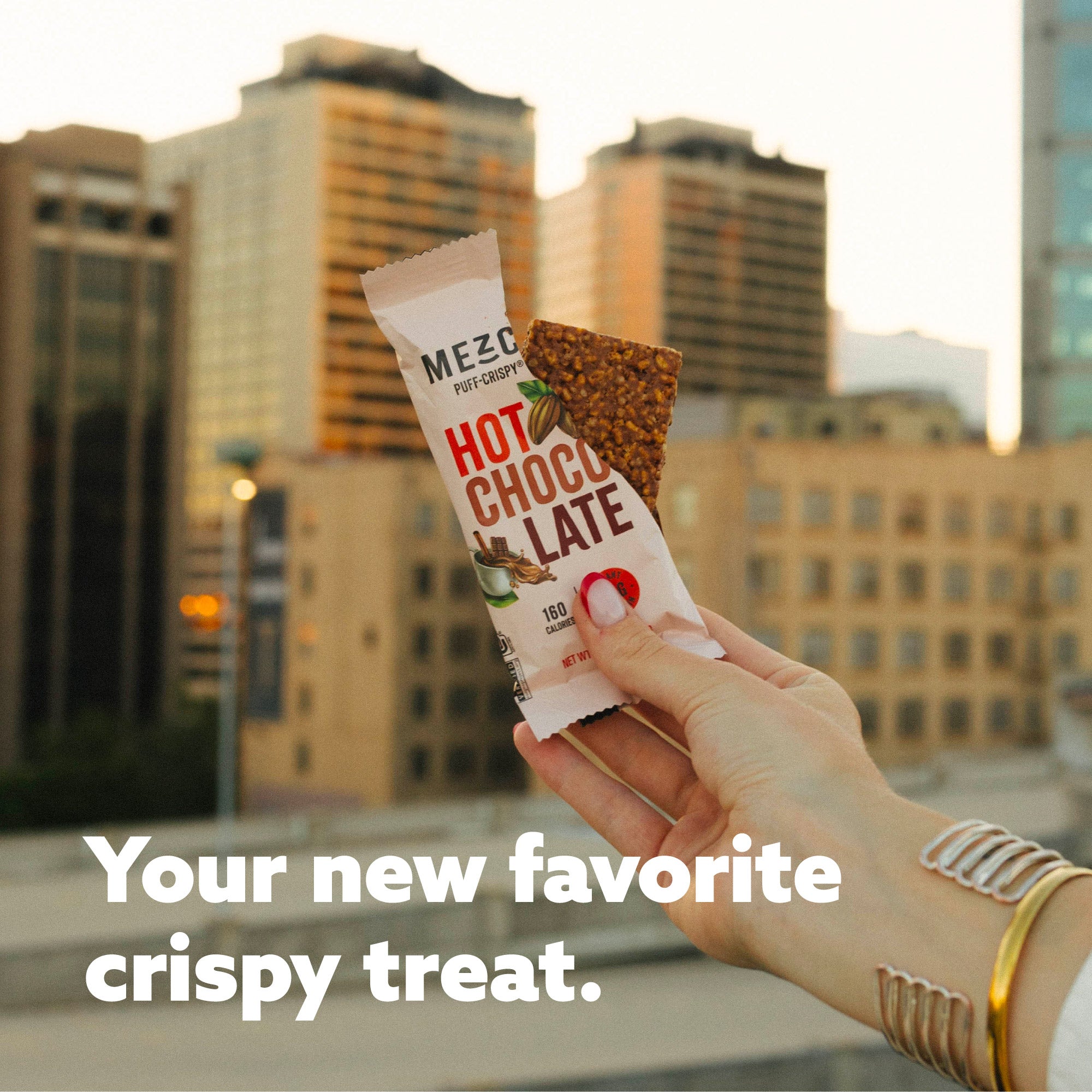 Hand holding MEZO Hot Chocolate puff-crispy snack bar against blurred city skyline background with marketing text