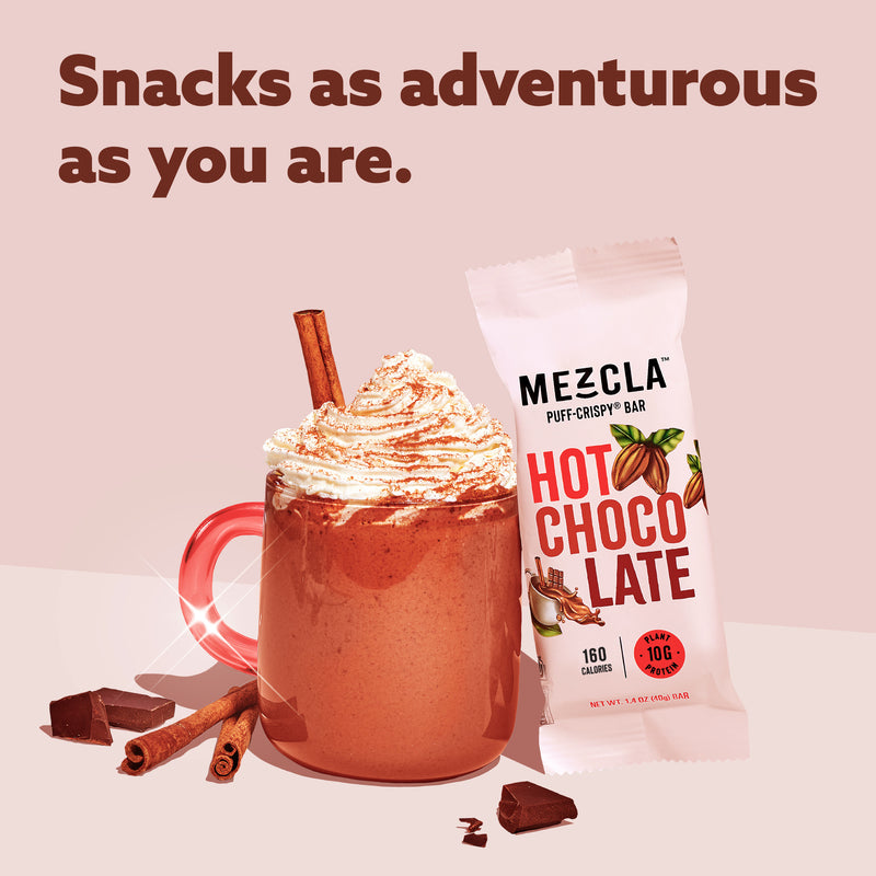 Pink background with red mug of hot chocolate topped with whipped cream and cinnamon stick, next to Mezcla Hot Chocolate protein bar package