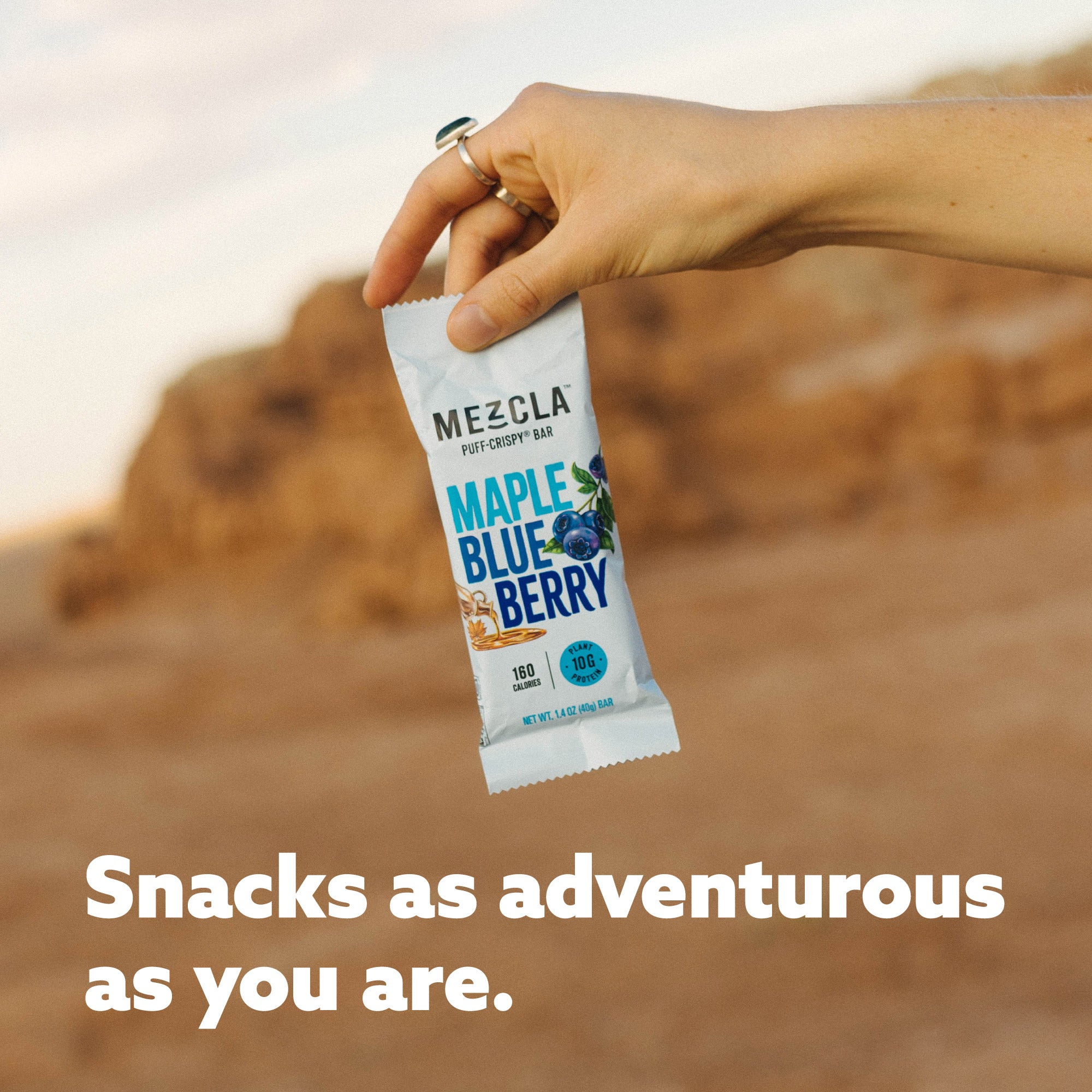 Hand holding Mezcla Maple Blueberry protein bar package against desert landscape with text "Snacks as adventurous as you are"