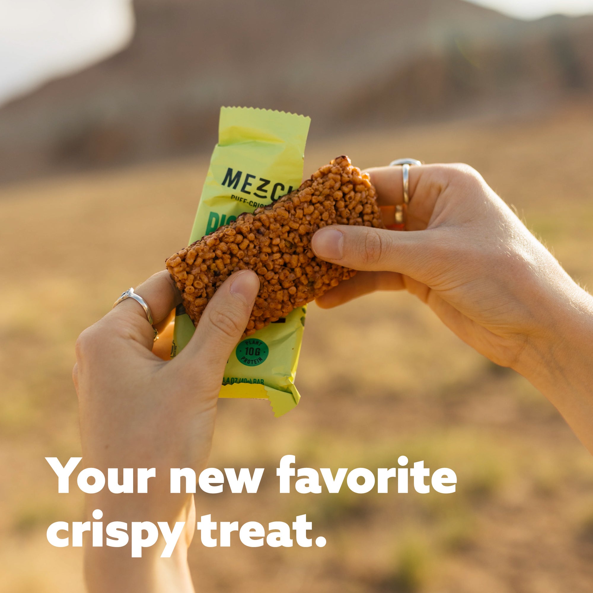 Hands holding a textured brown crispy bar from yellow Mezcla package against blurred outdoor background