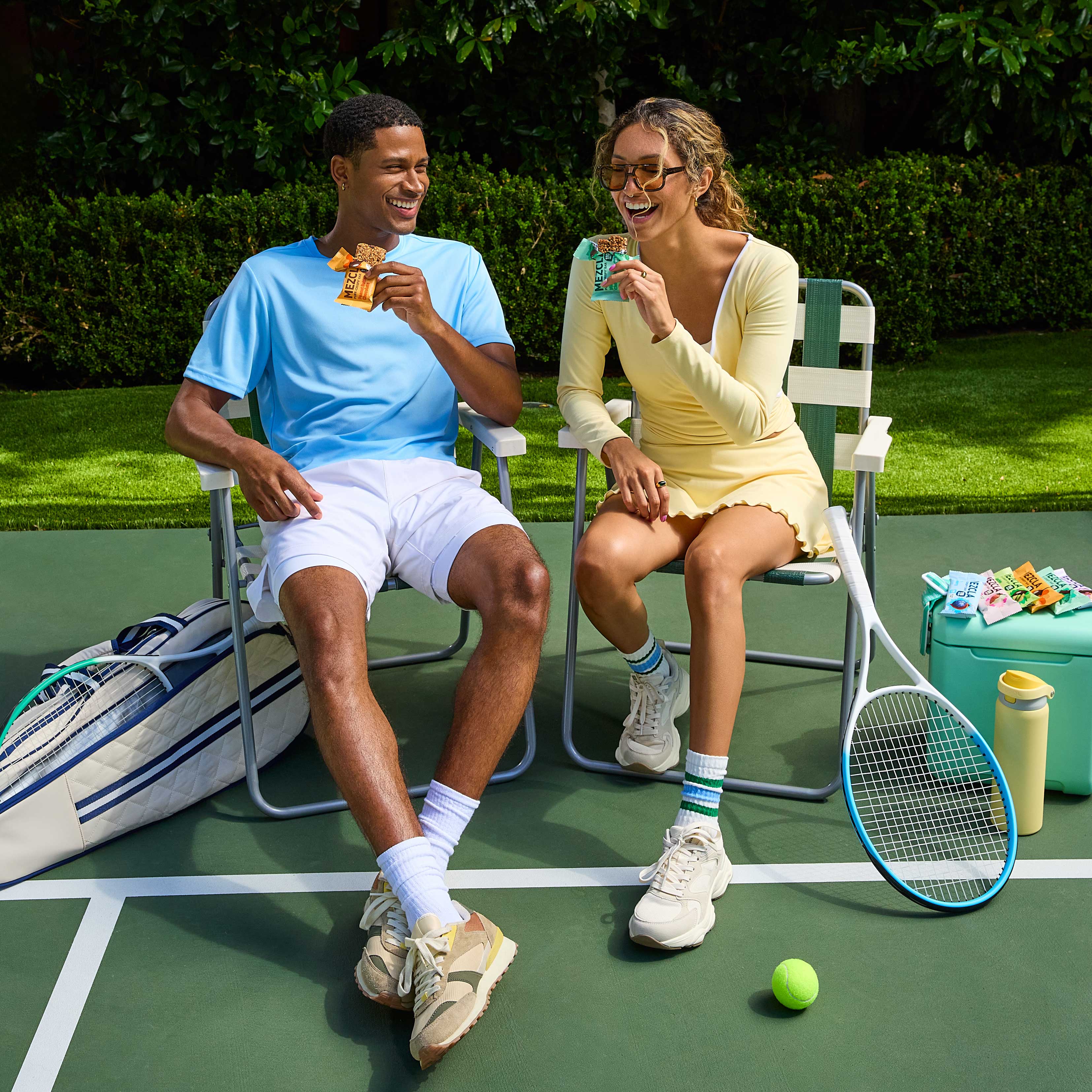 Two tennis players sitting on chairs, laughing while holding colorful snack bars on a green tennis court with rackets and gear nearby.