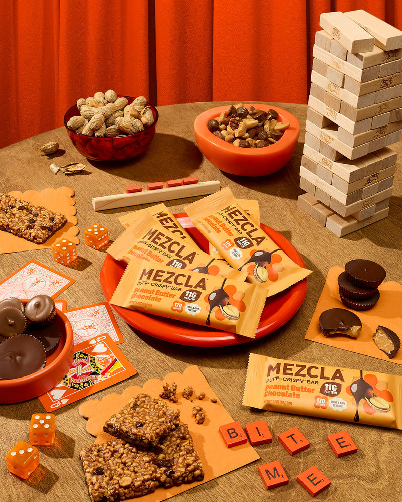 Orange-themed game night spread with Mezcla peanut butter chocolate protein bars, Jenga blocks, dice, playing cards, nuts, and chocolate treats on wooden table.