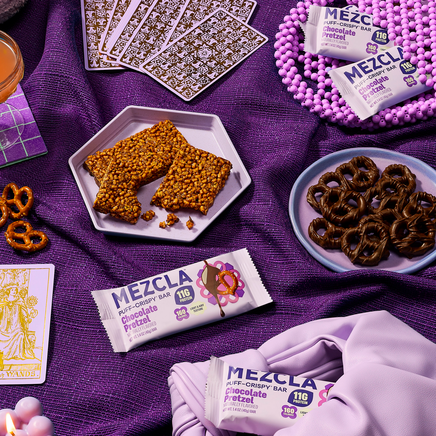 Purple-themed flat lay with MEZCLA Chocolate Pretzel protein bars, tarot cards, chocolate-covered pretzels, and granola bars on textured fabric