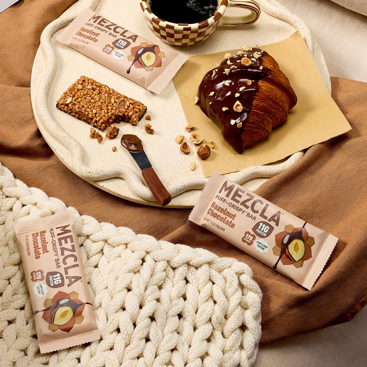 MEZCLA hazelnut chocolate protein bars on cream tray with chocolate croissant, coffee cup, and rope mat on brown fabric background