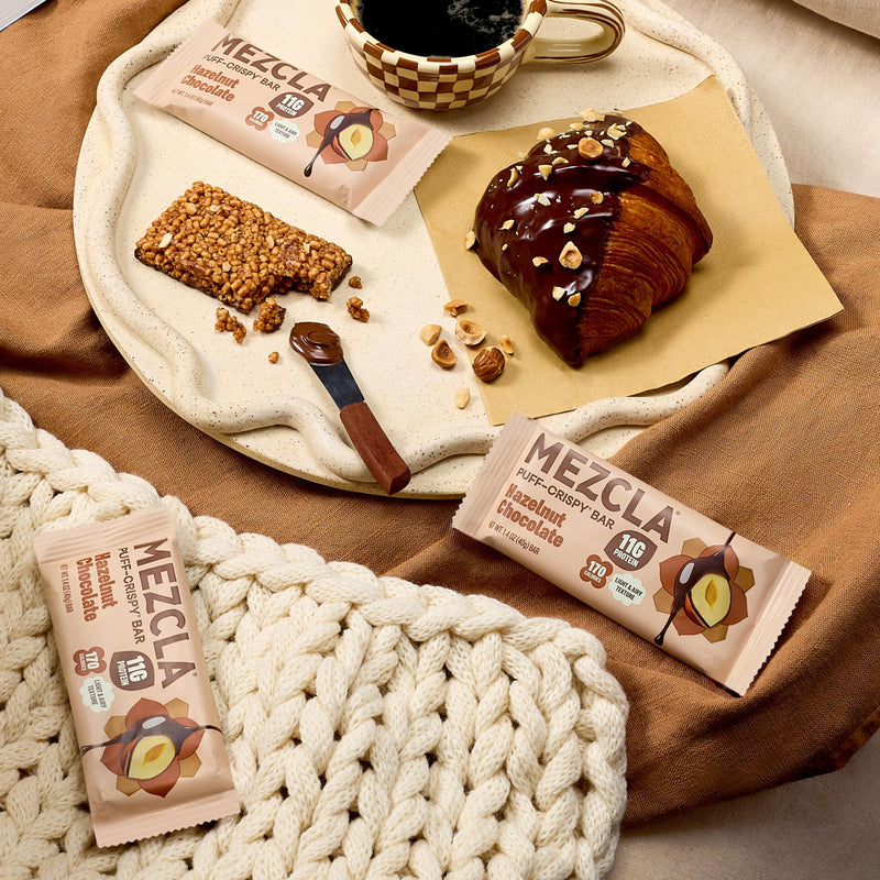 MEZCLA hazelnut chocolate protein bars on cream tray with chocolate croissant, coffee cup, and rope mat on brown fabric background