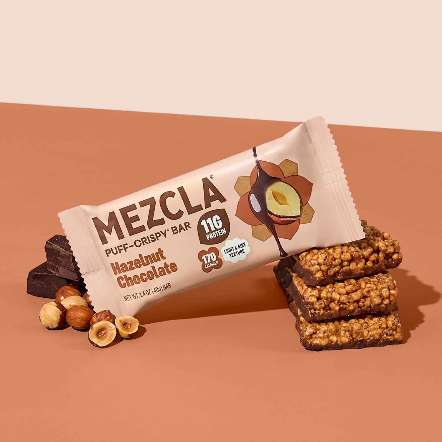 MEZCLA Hazelnut Chocolate puff-crispy protein bar package with 11g protein, 170 calories, shown with stacked bar pieces and hazelnuts on orange background