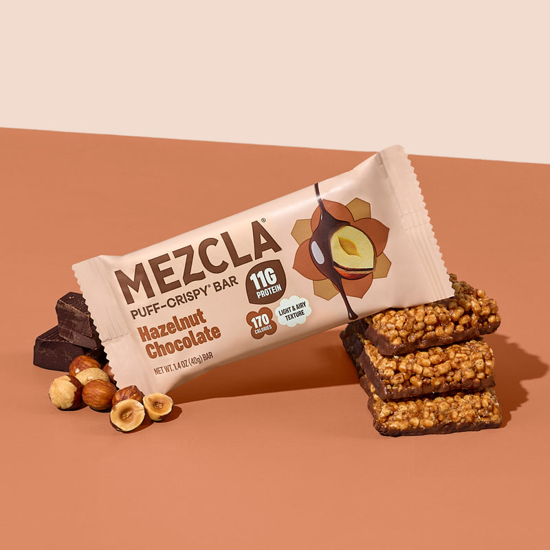 MEZCLA Hazelnut Chocolate puff-crispy protein bar package with 11g protein, 170 calories, shown with stacked bar pieces and hazelnuts on orange background
