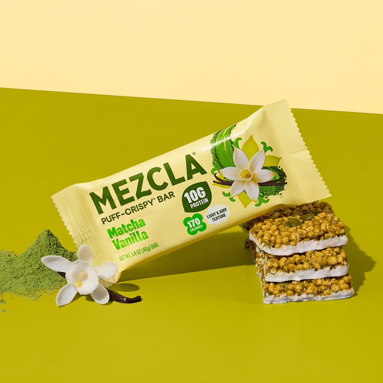 Mezcla Matcha Vanilla puff-crispy protein bar package on green surface with vanilla flowers and matcha powder, showing stacked bar pieces with white cream layers.
