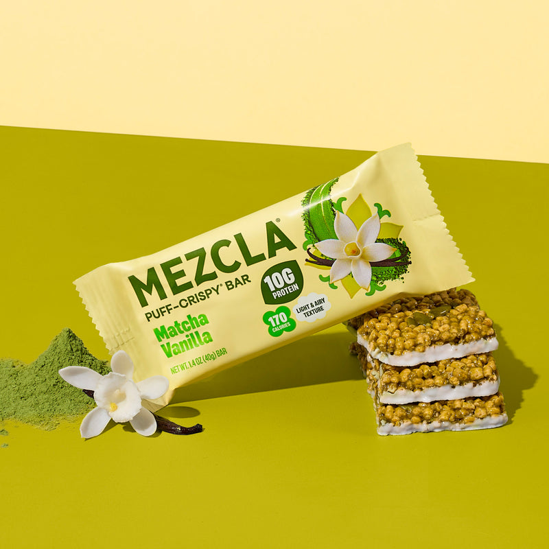 Mezcla Matcha Vanilla puff-crispy protein bar package on green surface with vanilla flowers and matcha powder, showing stacked bar pieces with white cream layers.