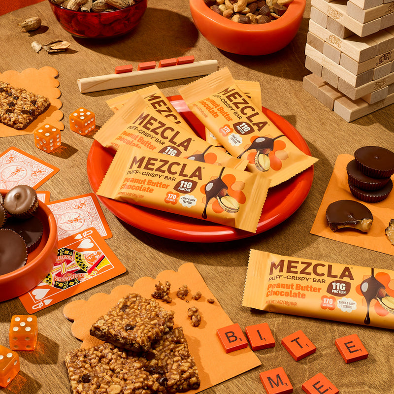MEZCLA peanut butter chocolate protein bars arranged on orange plate surrounded by games, nuts, and snacks on wooden surface