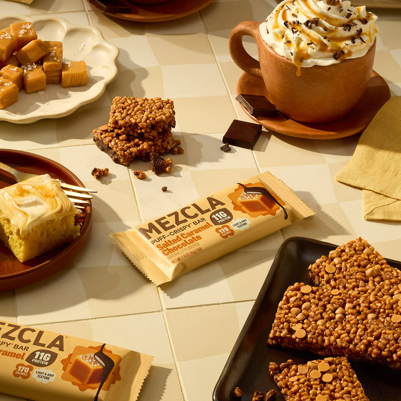 MEZCLA protein bars in salted caramel chocolate flavor displayed with coffee cup, caramel candies, and crispy treats on white tiles