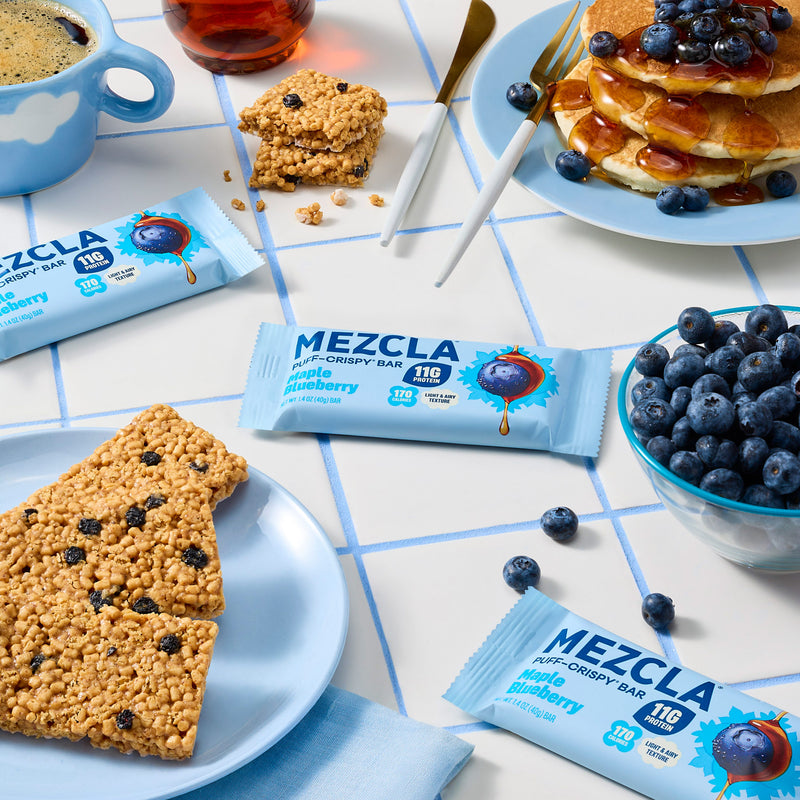 MEZCLA puff-crispy bars in maple blueberry flavor arranged on white tiled surface with fresh blueberries, pancakes, coffee, and granola bars