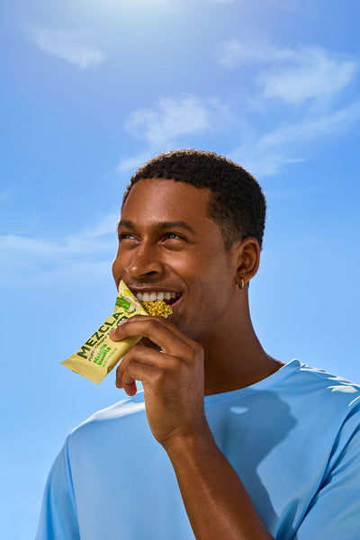 Young Black man in blue shirt smiling while eating a yellow Vega protein bar against bright blue sky with white clouds